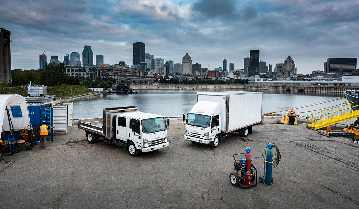 Photo of two Isuzu trucks by water with cityscape behind them.